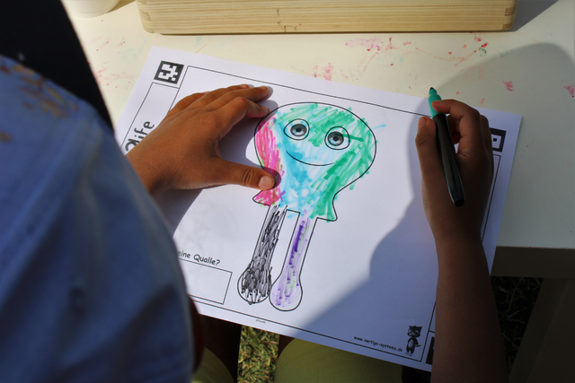 Ein Kind malt das Bild von einer Qualle mit bunten Farben an.