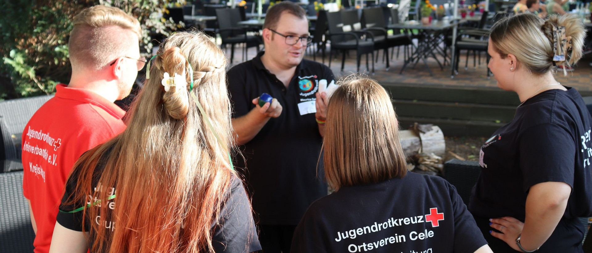 Mehrere Personen mit JRK-T-Shirts stehen im Kreis und lernen eine Aufgabe des Pflasterabzeichens kennen.