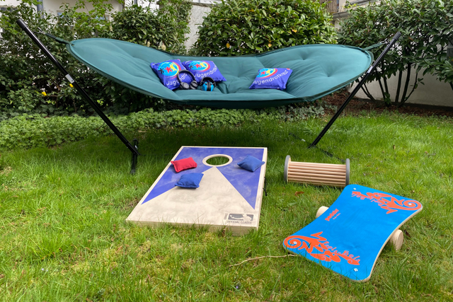 Eine Hängematte auf Standfüßen, ein Balanceboard, ein Pedalo und ein Wurfspiel liegen auf einer Wiese.