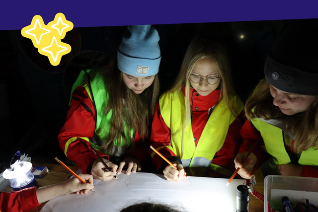 Nachtaufnahme von drei Personen, die mit Warnwesten bekleidet sind. Sie stehen um einen Tisch und arbeiten gemeinsam an einer Aufgabe.